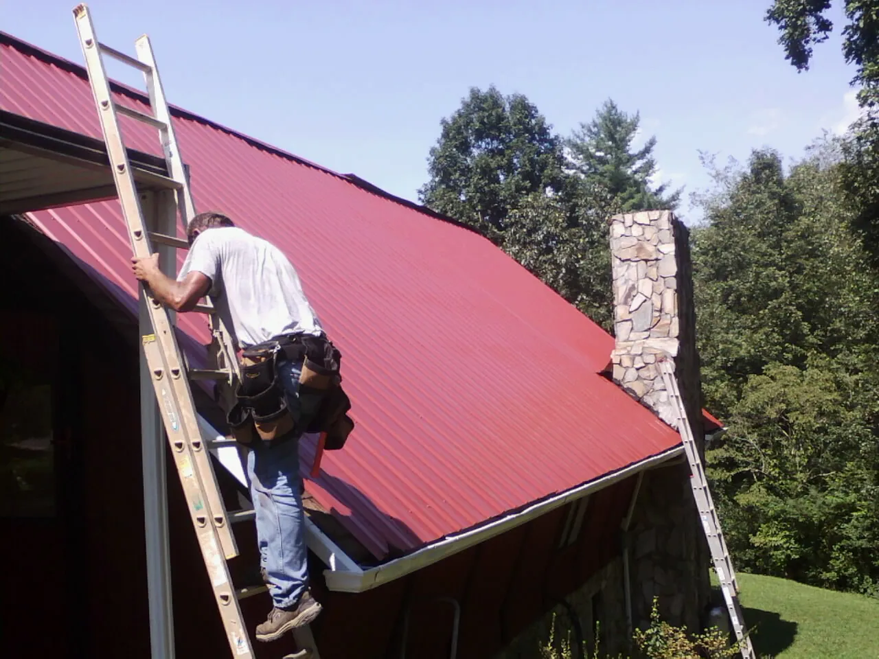 Roofer on a ladder beside a red metal roof and stone chimney — Soffit & Fascia Repair by Roofing Brothers in Newcastle